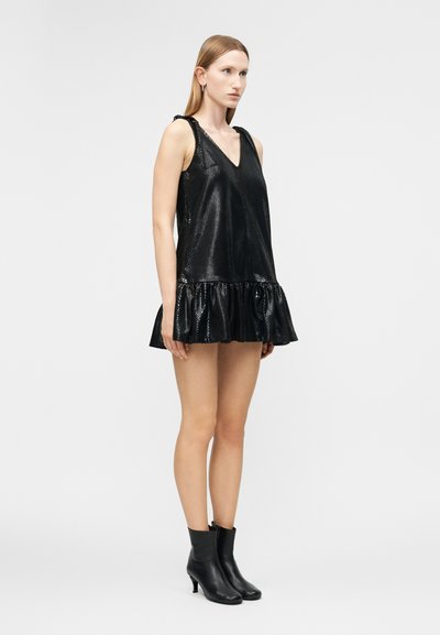 Kobieta stojąca w czarnej, błyszczącej, bezrękawnikowej mini sukience z falbaną na dole i czarnych, szpiczastych botkach na tle jasnego, jednolitego tła.