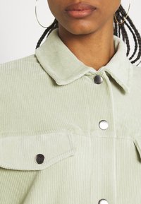 Giacca in corduroy verde chiaro con colletto, dotata di bottoni a pressione argentati e tasche sul petto, che presenta una texture a righe verticali.