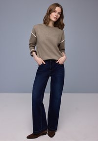 Femme aux cheveux bruns mi-longs portant un pull beige, un jean large bleu foncé et des chaussures marron, debout les mains dans les poches.