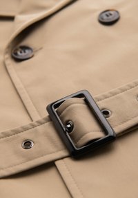 Tissu de trench-coat beige avec boucle de ceinture rectangulaire noire, ceinture cousue et boutons foncés avec surpiqûres visibles des boutonnières.