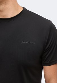 Κοντινό πλάνο ενός ατόμου που φοράει μαύρο T-shirt Calvin Klein με κοντά μανίκια και στρογγυλή λαιμόκοψη.