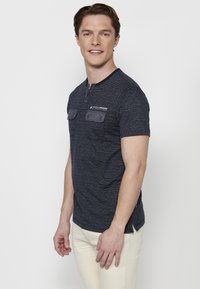 Tumši zilas krāsas īssvārku krekls ar teksturētu rakstu, diviem krūšu kabatām un henley tipa apkakli. Iezīmēta ar diskrētu zīmolu un sānu šķēlumiem.