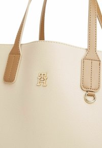 Beżowa skórzana torba na ramię o teksturowanym wykończeniu, z jasnobrązowymi uchwytami, złotym akcentem logo oraz metalowym detalem w postaci pierścienia.