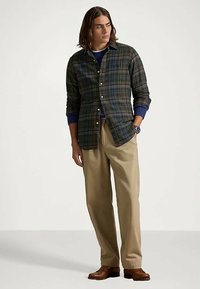 Mlad moški z dolgimi lasmi, oblečen v temno karirano srajco s kratkimi rokavi, modro jopico, bež hlače, rjave čevlje in uro na zapestju, stoji z eno roko v žepu.