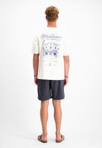 Witte t-shirt met korte mouwen en een blauwe grafische print op de achterkant, die een gebouw en vogels afbeeldt. Gekleed met gestructureerde donkere shorts.