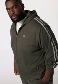 Mørkeolivengrøn zip-up hoodie med en tætsiddende design, der har grå striber på ærmerne og sidelommer. Tekstureret stof med logodetaljer.