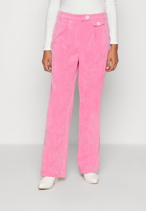 Woman wearing high-waist, wide-leg pink corduroy pants with button details paired with white long-sleeve top and white shoes.