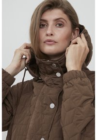 Brun quilteret jakke med høj krave og hætte, der har knaplukninger og snørede ærmer, lavet af blødt, tekstureret stof.