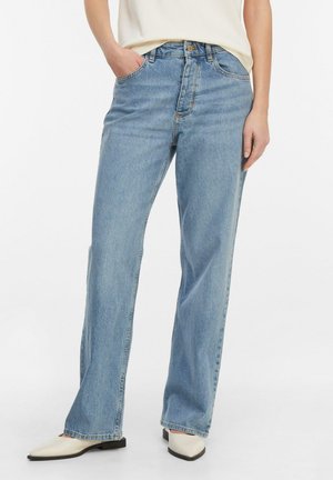 Kobieta ubrana w jasnoniebieskie dżinsy o prostych nogawkach, białe buty z ostrym noskiem oraz wpuszczoną do środka kremową bluzkę, stojąca z jedną ręką w kieszeni.