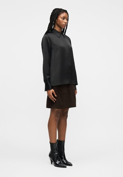 Γυναίκα με μακριά πλεξούδες, φοράει μαύρο σατινέ μπλουζάκι με μακριά μανίκια, σκουρό καφέ φούστα μέχρι το γόνατο με μπροστινή τσέπη και μαύρα παπούτσια με τακούνι από δερμάτινο λάκκο.