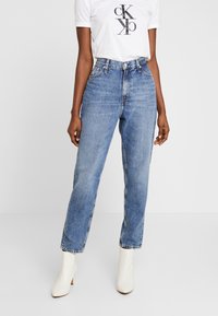 Kvinna som bär högmidjade blå jeans, vita ankelstövlar och en vit t-shirt med svart Calvin Klein-logotyp mot en enfärgad bakgrund.