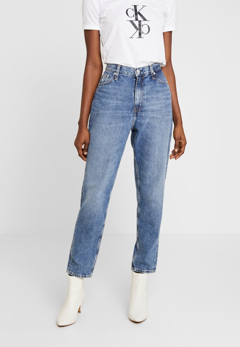 Kvinna som bär högmidjade blå jeans, vita ankelstövlar och en vit t-shirt med svart Calvin Klein-logotyp mot en enfärgad bakgrund.