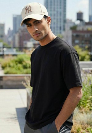 Junger Mann mit beiger Baseballkappe und schwarzem T-Shirt, der im Freien steht, im Hintergrund städtische Gebäude.
