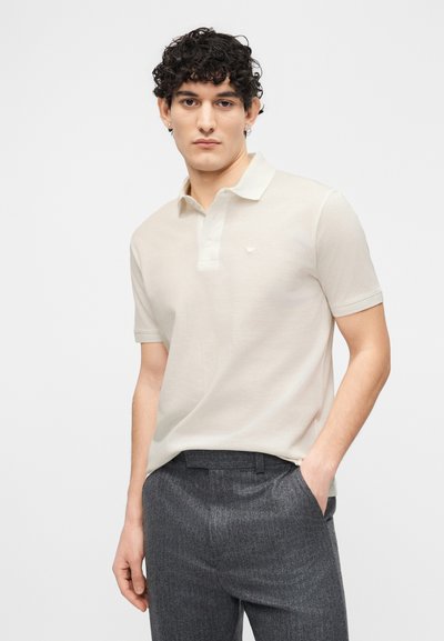 Νεαρός με σγουρά σκούρα μαλλιά φοράει μπεζ πόλο με κοντά μανίκια και γκρι ραμμένα παντελόνια, στέκεται μπροστά σε απλό λευκό φόντο.