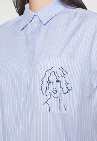 Chemise bleu clair boutonnée avec fines rayures blanches et une petite poche ornée d'un dessin au trait noir brodé représentant un visage de femme.