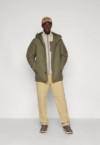 Alyvuogių žalios spalvos pūkinė striukė su gaubtu, smėlio spalvos tekstūruotas megztinis, smėlio spalvos kelnės ir tamsūs sportbačiai, derinami su ruda kepure ir laisvu stovėjimu.