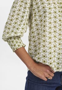 Blusa bianca con ricami floreali verdi. Maniche a sbuffo con polsini arricciati. Tessuto liscio. Indossata con pantaloni in denim blu.