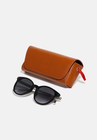 Des lunettes de soleil noires en forme d'œil de chat avec des verres dégradés, accompagnées d'un étui en cuir marron doté d'un tirant rouge et d'un logo en relief.