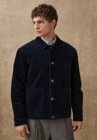 Giacca in velluto a coste blu scuro con colletto, chiusura frontale con bottoni, due tasche a toppa e tessuto testurizzato; indossata sopra una camicia azzurro chiaro.