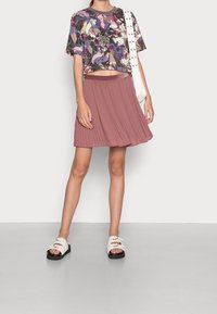 Kortärmad topp med en flerfärgad blommönster, kombinerad med en plisserad rosaskjol och vita sandaler med svarta sulor.