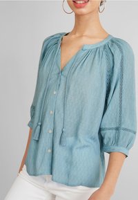Blouse bleu clair en tissu texturé, avec un col en V, des boutons sur le devant, des pompons et des accents de dentelle sur les manches. Manches bouffantes trois-quarts.