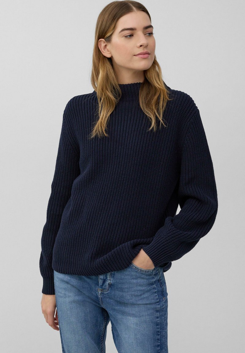 Marineblauwe geribbelde gebreide trui met een hoge hals, lange mouwen en een ontspannen pasvorm, gecombineerd met blauwe jeans, weergegeven tegen een lichtgrijze achtergrond.