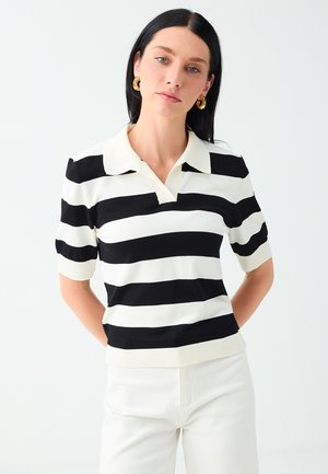Donna con capelli neri indossa una maglietta a maniche corte a righe orizzontali bianche e nere e pantaloni bianchi su sfondo chiaro uniforme.