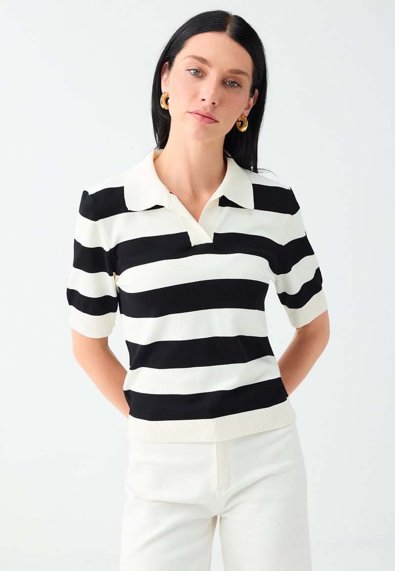Femme aux cheveux noirs portant un t-shirt à manches courtes rayé horizontalement noir et blanc et un pantalon blanc sur un fond clair uni.