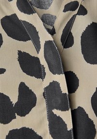 Kravata s leopardím potiskem v béžové barvě s velkými černými nepravidelnými skvrnami. Má design s hlubokým výstřihem a viditelným švem kolem okrajů.