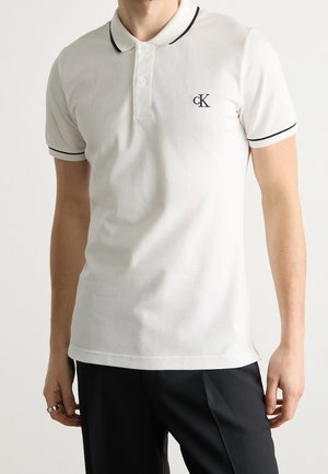 Homme portant un polo blanc avec bordure noire et logo cK, associé à un pantalon noir, debout devant un fond uni.