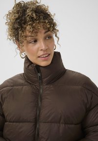 Ruda pūkine striukė su aukštu apykakle, užtrauktuko detalėmis, išklotomis struktūromis ir lengvu medžiagų pasirinkimu. Modelis turi garbanotus plaukus ir dėvi auksines kilpines auskarus.