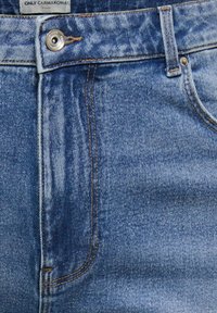 Modré džínsy z denimu v detailnom zábere, ukazujúci predný vrecko, pútk na opasok, kovové gombík, detaily šitia a značkovú štítok "ONLY CARMAKOMA" na páse.