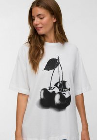 Vrouw met lang bruin haar die een oversized wit T-shirt draagt met een grote zwart-witte print van drie kersen met een blad.