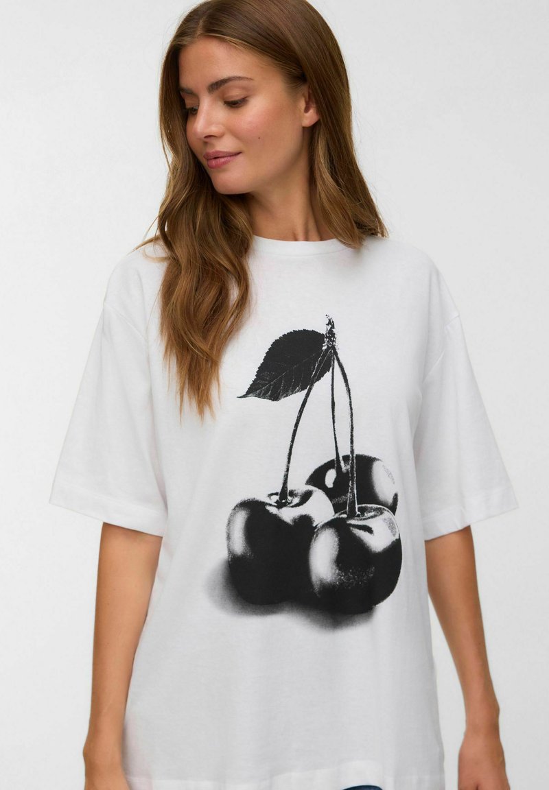 Vrouw met lang bruin haar die een oversized wit T-shirt draagt met een grote zwart-witte print van drie kersen met een blad.