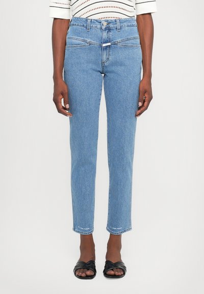 Jeans bleus taille haute avec une coupe droite, présentant un design épuré, un léger délavage et un ourlet effiloché. Portés avec des sandales noires.