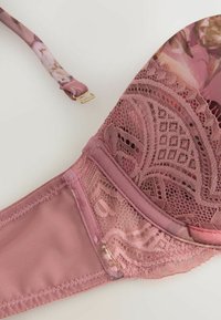 Close-up of mauve lace bra cup and strap with floral pattern and gold hook on white background.