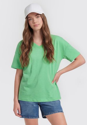 Jonge vrouw met lang bruin haar draagt een witte pet, een groen T-shirt met korte mouwen en V-hals, en blauwe denim shorts, staand tegen een effen achtergrond.