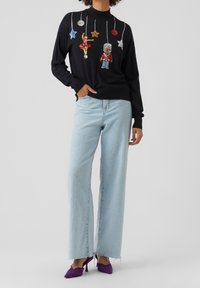 Pull noir avec des décorations colorées en sequins représentant des ornements et un casse-noisette. Associé à un jean large bleu clair et des talons violets.