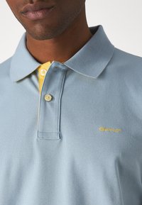 Polo in cotone azzurro chiaro con dettagli gialli nel colletto, placca con tre bottoni e logo "GANT" ricamato in giallo. Tessuto testurizzato.
