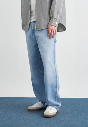 Asmuo dėvi šviesiai mėlynas platėjusias džinsines kelnes, baltus sportinius batelius su smėlio spalvos akcentais ir dryžuotą marškinius su rankovėmis nuleistomis.