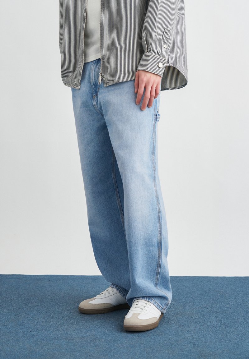 Άτομο που φοράει ανοιχτό μπλε τζιν παντελόνι με φαρδιά πόδια, λευκά αθλητικά παπούτσια με μπεζ λεπτομέρειες και ριγέ πουκάμισο με κουμπιά και μακριά μανίκια.