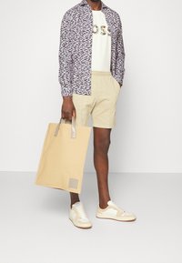 Model trägt ein gemustertes Hemd, ein weißes T-Shirt mit Text, beige Shorts, cremefarbene Sneakers und hält eine beige Tote-Bag mit grauen Akzenten.