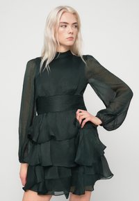 Νεαρή γυναίκα με μακριά ξανθά μαλλιά, φορώντας ένα διάφανο, μακρυμάνικο μαύρο φόρεμα με στρωτές ρίγες και ζώνη ψηλής μέσης, κοιτάζοντας πλάγια.