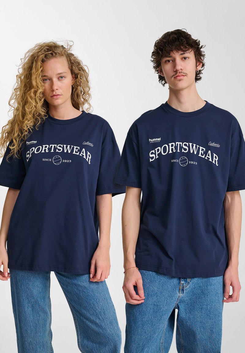 Marinblå bomulls t-shirts med kort ärm och rund hals, med vit text och logotyp "SPORTSWEAR". Bärs med ljusblå denimjeans.