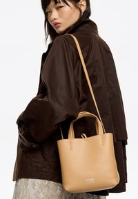 Borsa in pelle tan con doppi manici, texture liscia, design minimalista e logo del marchio impresso. Indossata con un giubbotto marrone scuro.