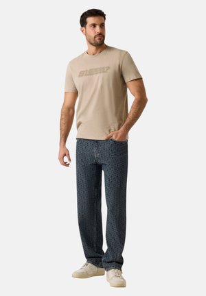 Uomo in piedi con una mano in tasca, indossa una t-shirt beige con logo, jeans blu scuro a motivo e scarpe da ginnastica bianche su uno sfondo semplice.