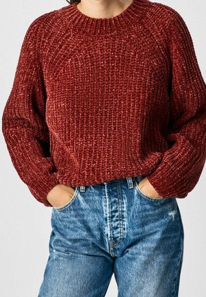 Persoon draagt een grofgebreide rode trui met lange mouwen en heeft de handen in de zakken van lichtblauwe spijkerbroeken gestoken.