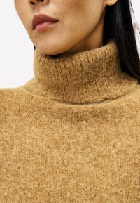 Kameelkleurige coltrui gemaakt van gestructureerde wol met een geribbelde kraag en een zachte, pluizige uitstraling. Close-up weergave van de stof.