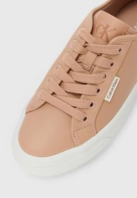Beige Calvin Klein leather sneaker with matching laces, white sole, and visible Calvin Klein logo tag on side.