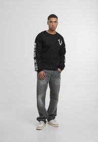 Black sweatshirt with logo on chest and white text on sleeves, paired with faded gray jeans and white sneakers. Standing against plain background.